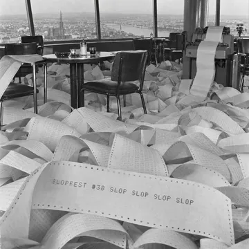 Photo of teleprinter paper piled knee-deep on a revolving café floor, the top sheet reading 'SLOPFEST #38 SLOP SLOP SLOP SLOP'